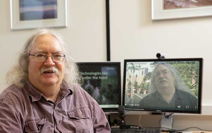 White haired distinguished man seated at office desk, on the computer screen behind is an image of the same man many years earlier speaking in an interview